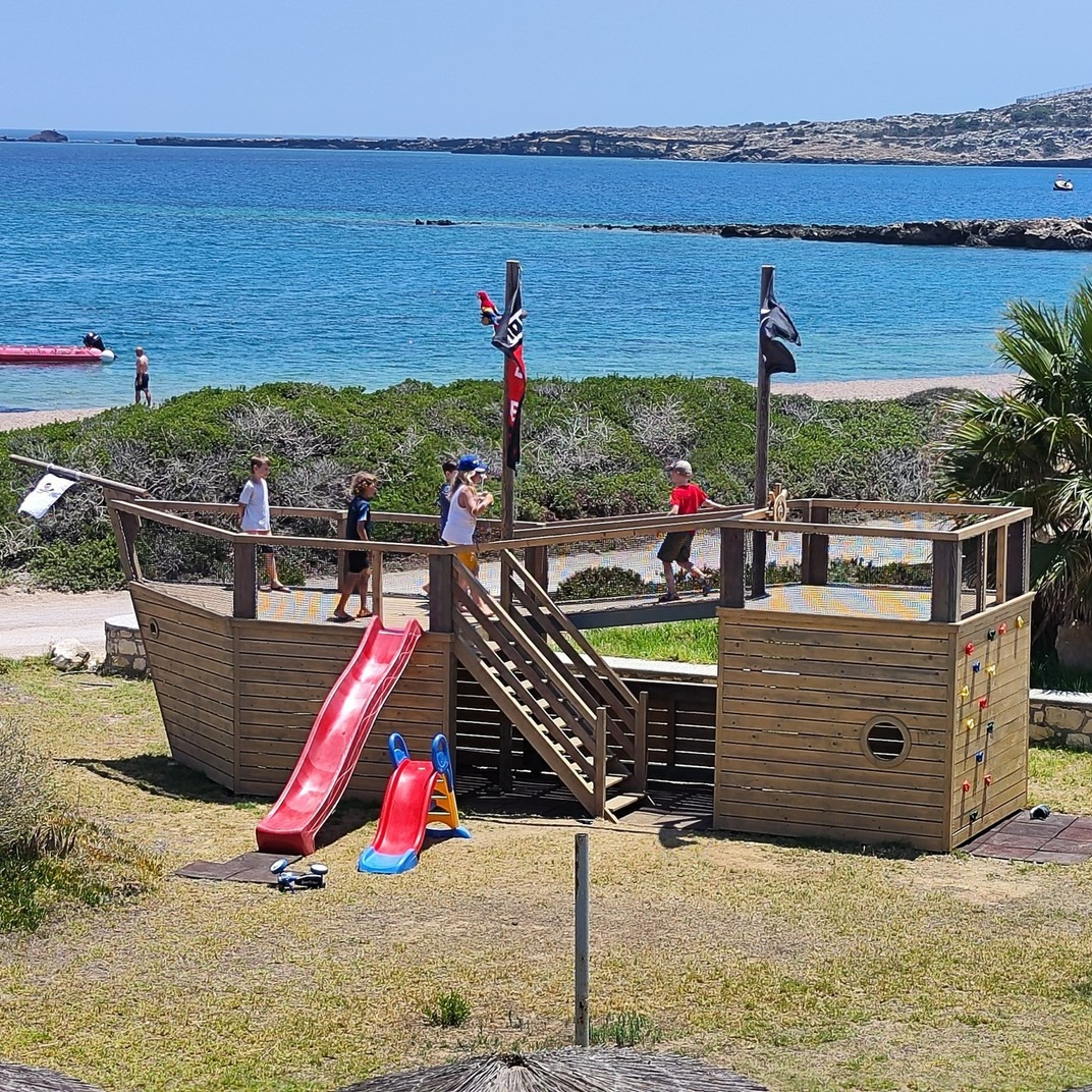 Family corner of the bay — Pirate-ship play structure on the beach. Knee-deep water steps away. Kids supervised, parents on the foil.
