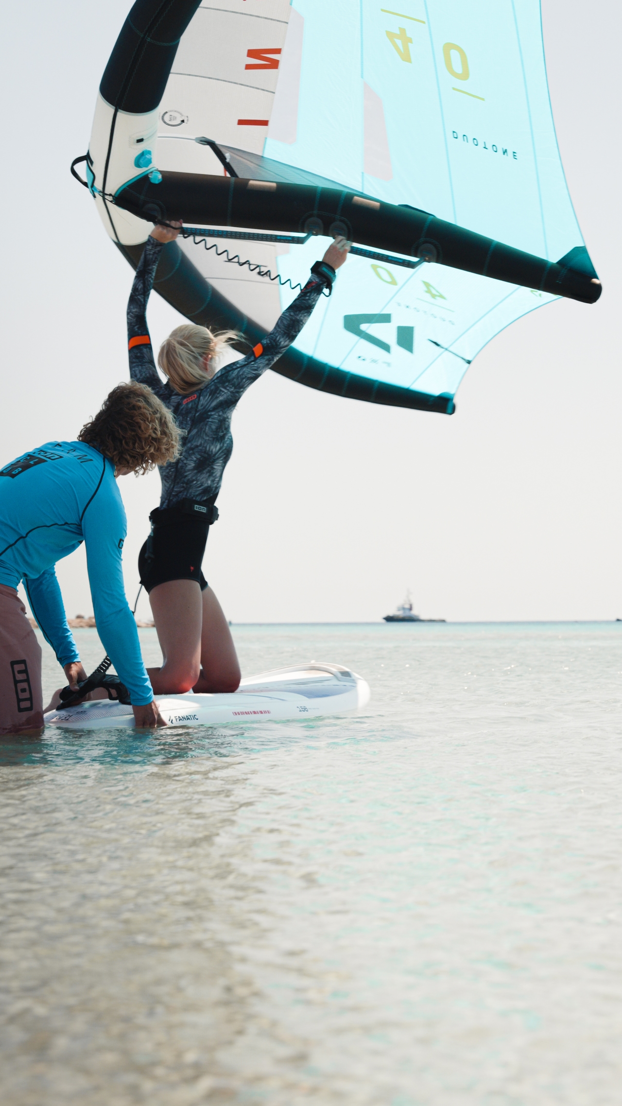Coach guiding a first-time wingfoiler kneeling on the board in knee-deep water at ION Karpathos